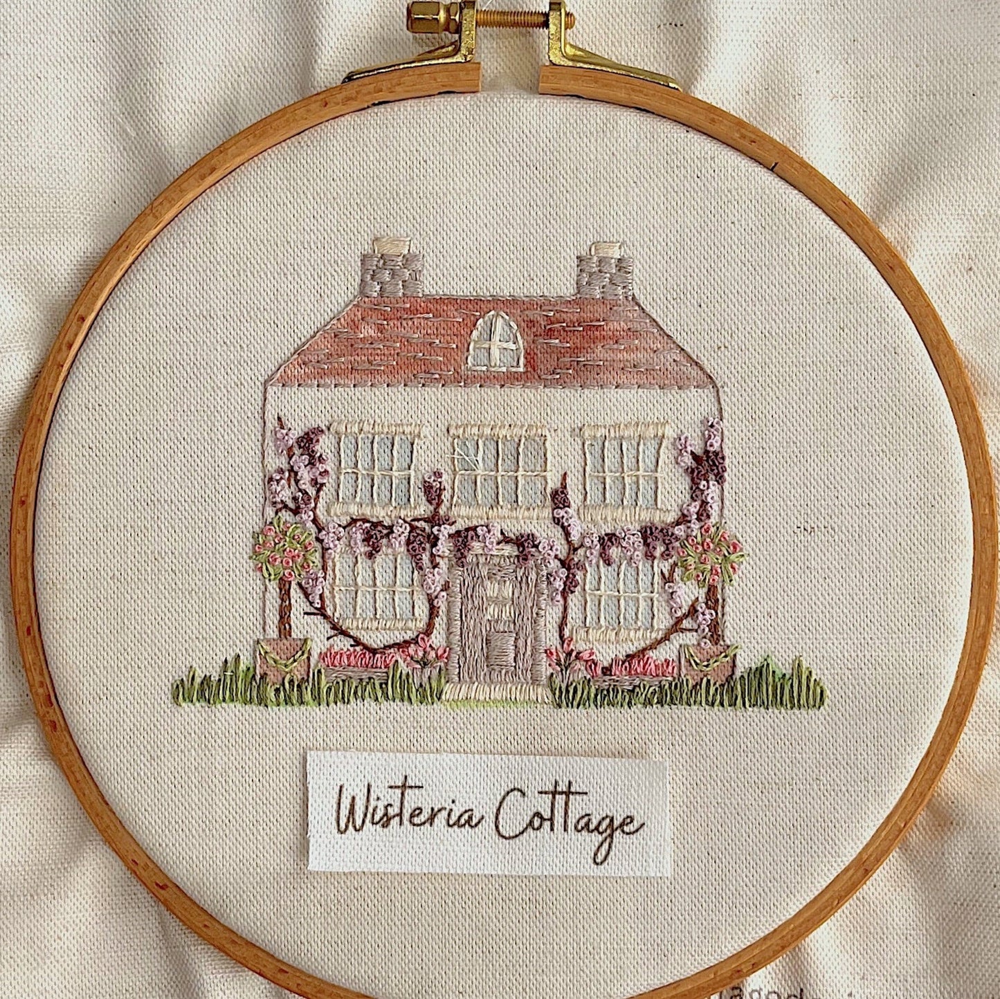 Embroidery of a house with wisteria vines in a wooden hoop on beige fabric.
