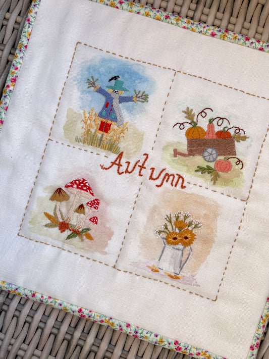 A square white fabric textile featuring four embroidered autumn-themed scenes: a scarecrow, pumpkins in a wagon, mushrooms, and sunflowers, with the word 'Autumn' stitched in the center. The fabric has a floral border and is laid on a woven wicker surface.