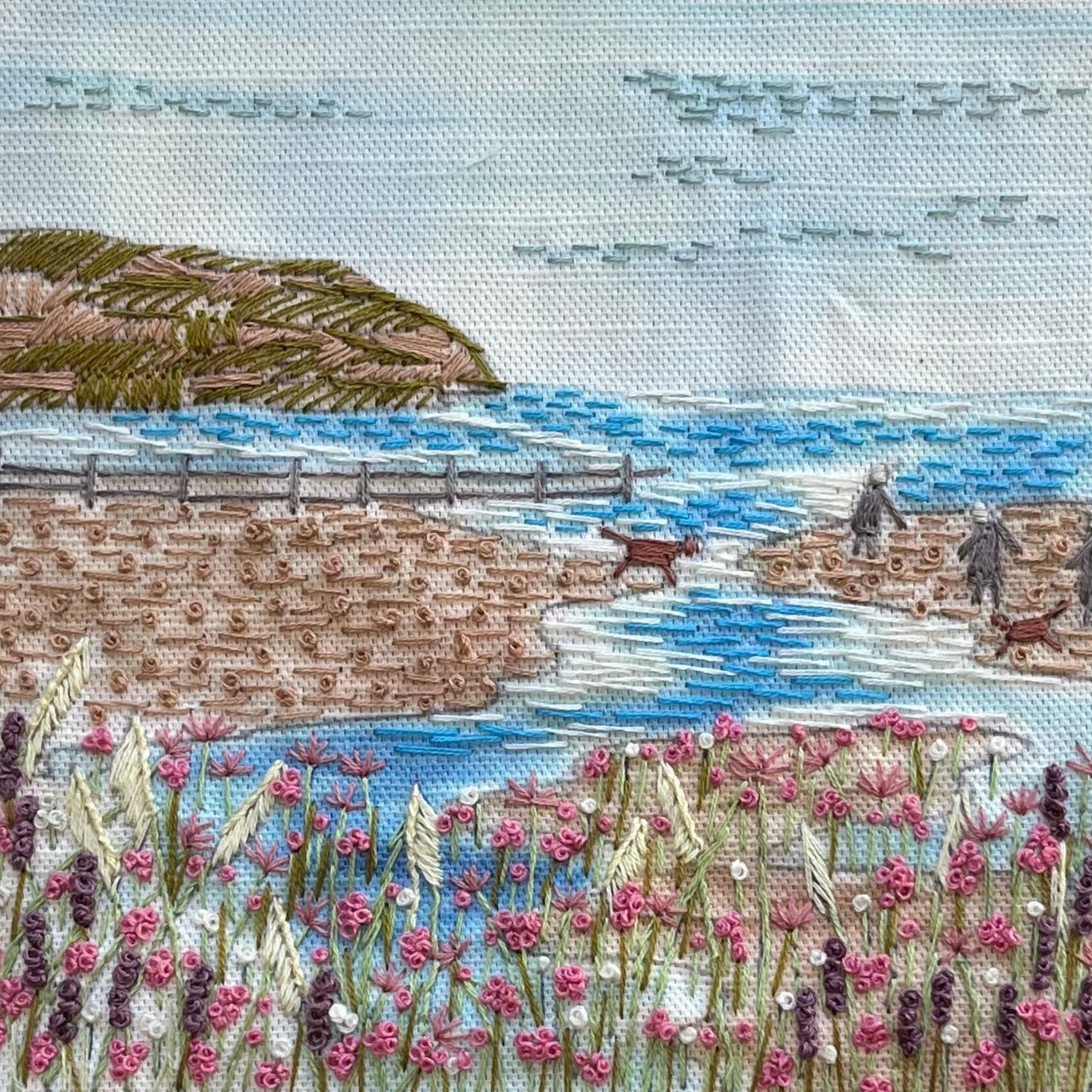 Embroidered scene of a coastal landscape with a wooden pier, dog, and people.
