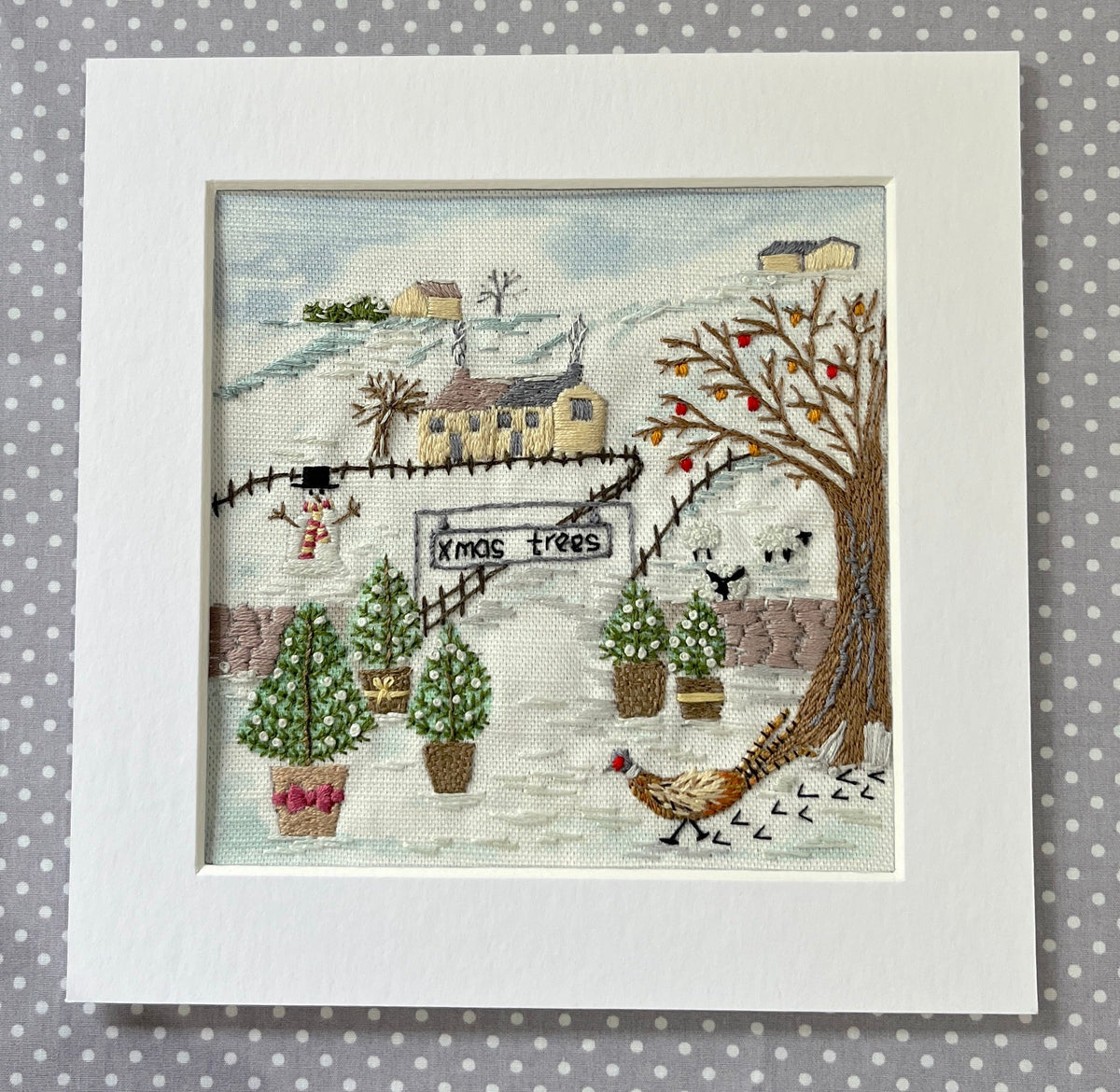 A finished cross-stitch embroidery piece featuring a winter village scene with potted Christmas trees, a sign reading 'MOS trees', houses, birds, and a pheasant, presented in a white square mat against a gray polka-dot background.
