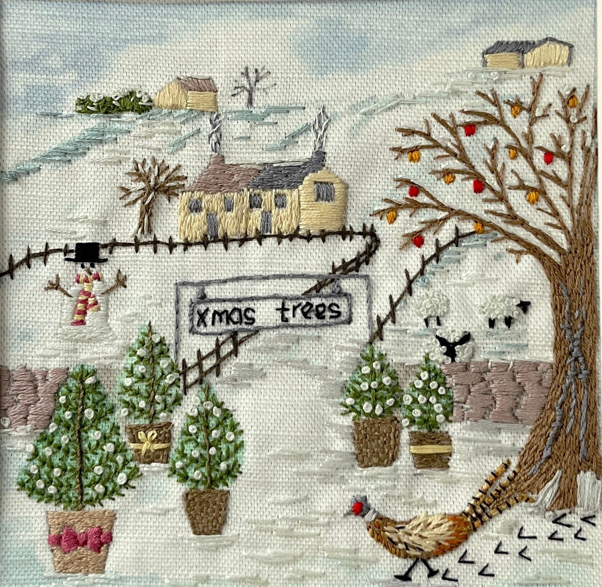 A finished cross-stitch embroidery piece featuring a winter village scene with potted Christmas trees, a sign reading 'MOS trees', houses, birds, and a pheasant, presented in a white square mat against a gray polka-dot background.