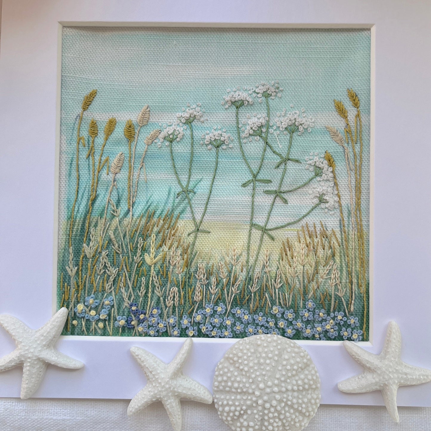 embroidery of a natural scene with plants and flowers, displayed on a white surface with starfish and a shell.