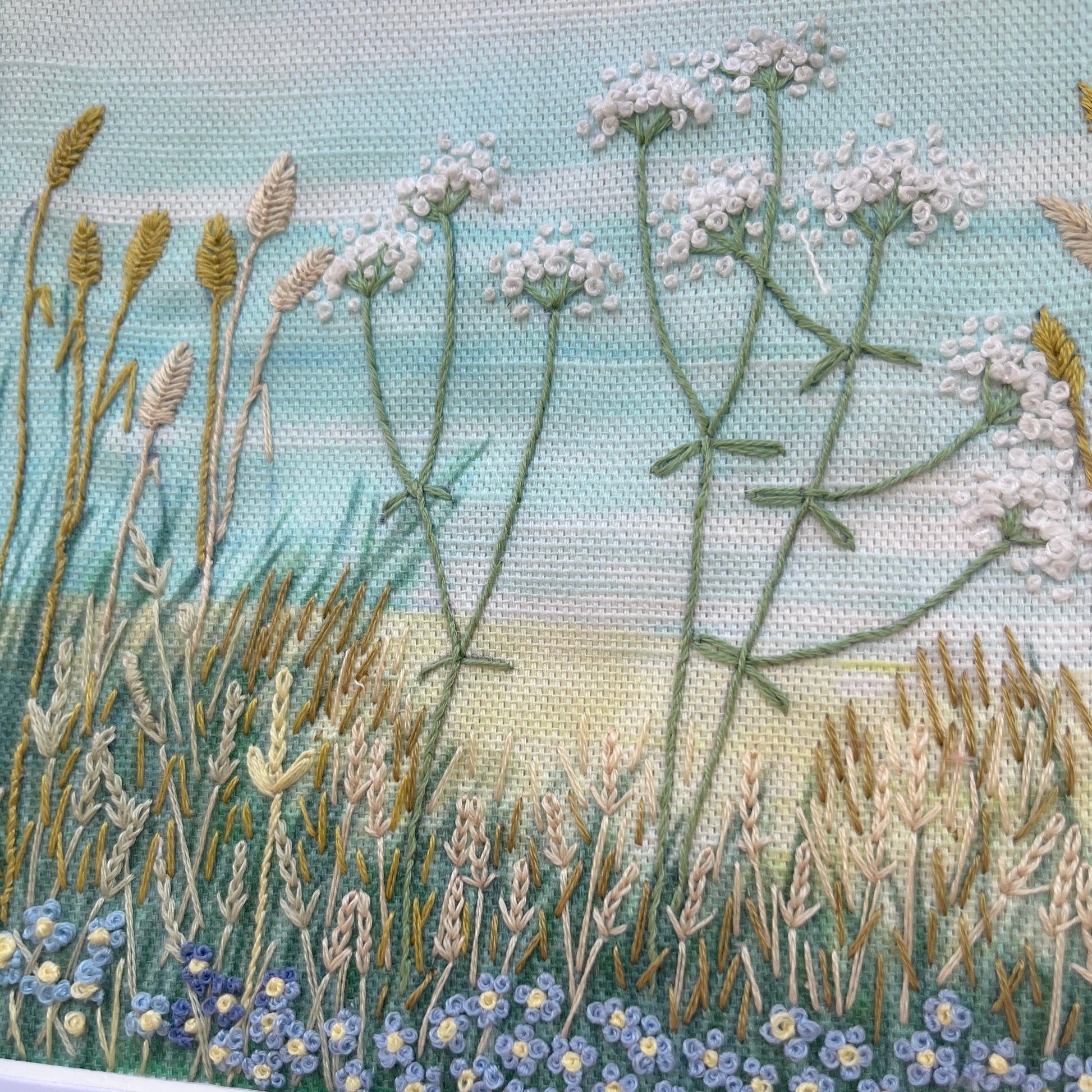 Embroidered scene of a field with flowers and grasses on a light blue background.