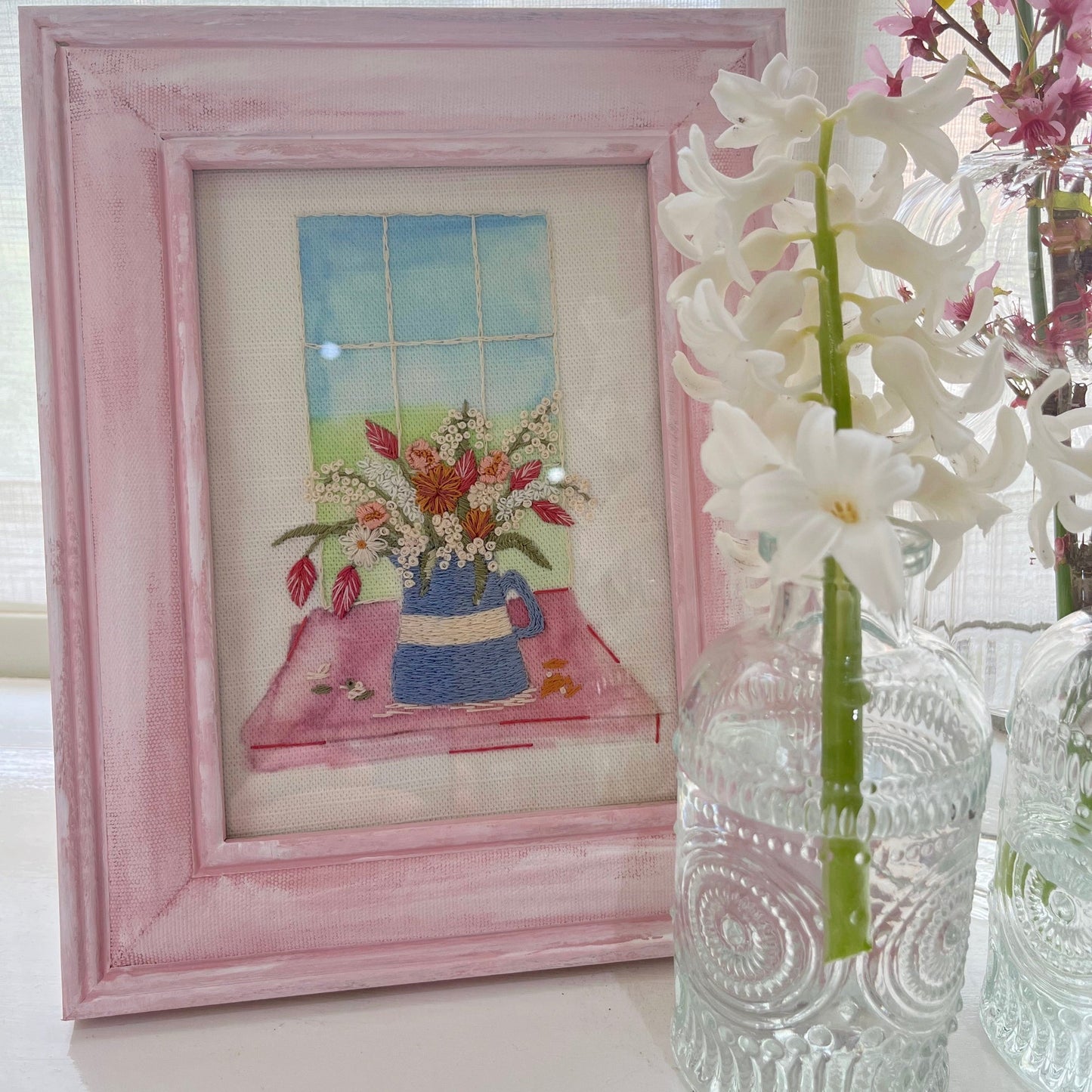 Framed artwork with a pink border next to a clear glass vase with white flowers.