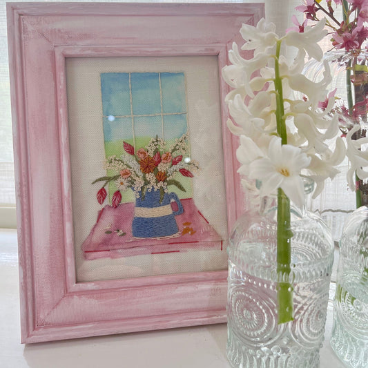 Framed artwork with a pink border next to a clear glass vase with white flowers.
