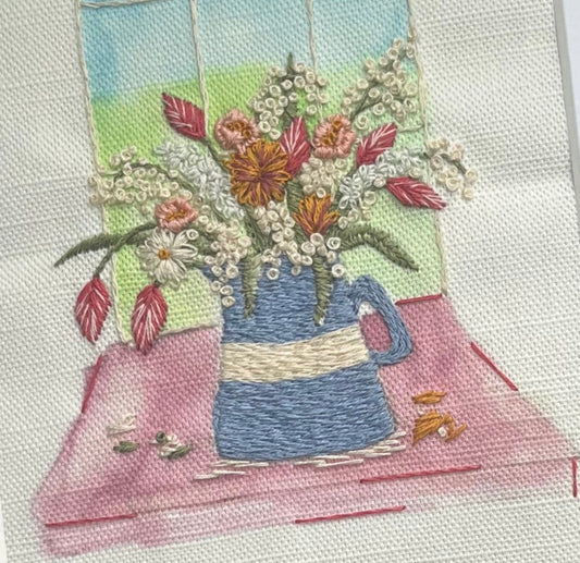Embroidery of a blue teapot with flowers on a pink tablecloth, framed by a window view.