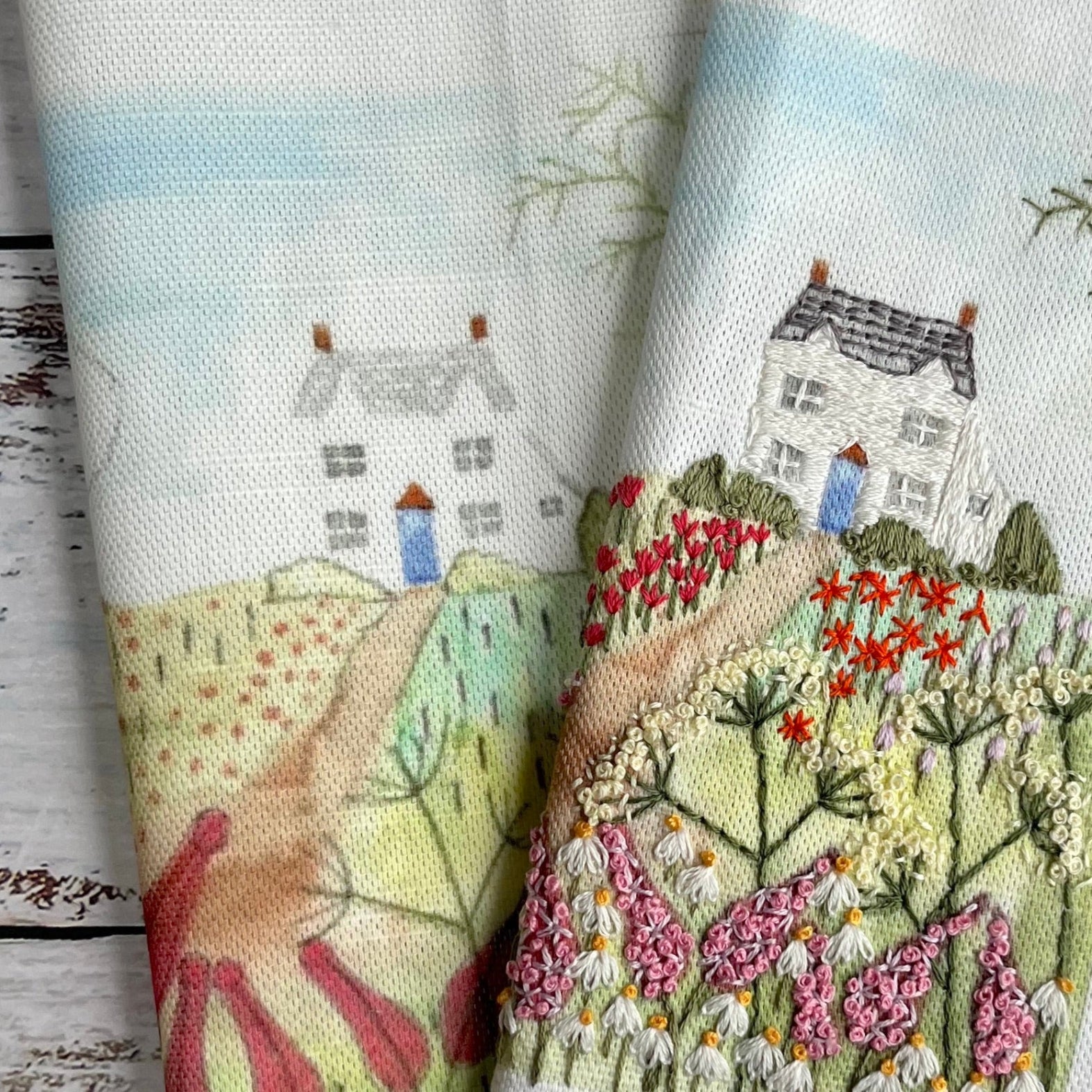 Embroidered fabric with a scenic design of houses and gardens on a wooden surface.