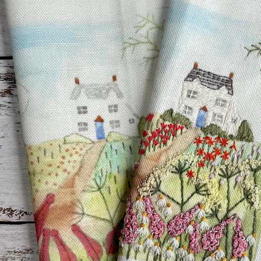 Embroidered fabric with a scenic design of houses and gardens on a wooden surface.