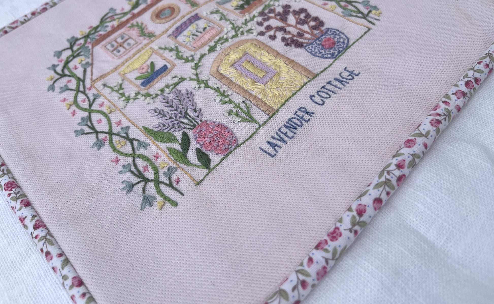 A square embroidered textile featuring a cottage design with the words 'LAVENDER COTTAGE' and floral vine motifs, finished with a pink and white floral piped border, laid on a white fabric background.