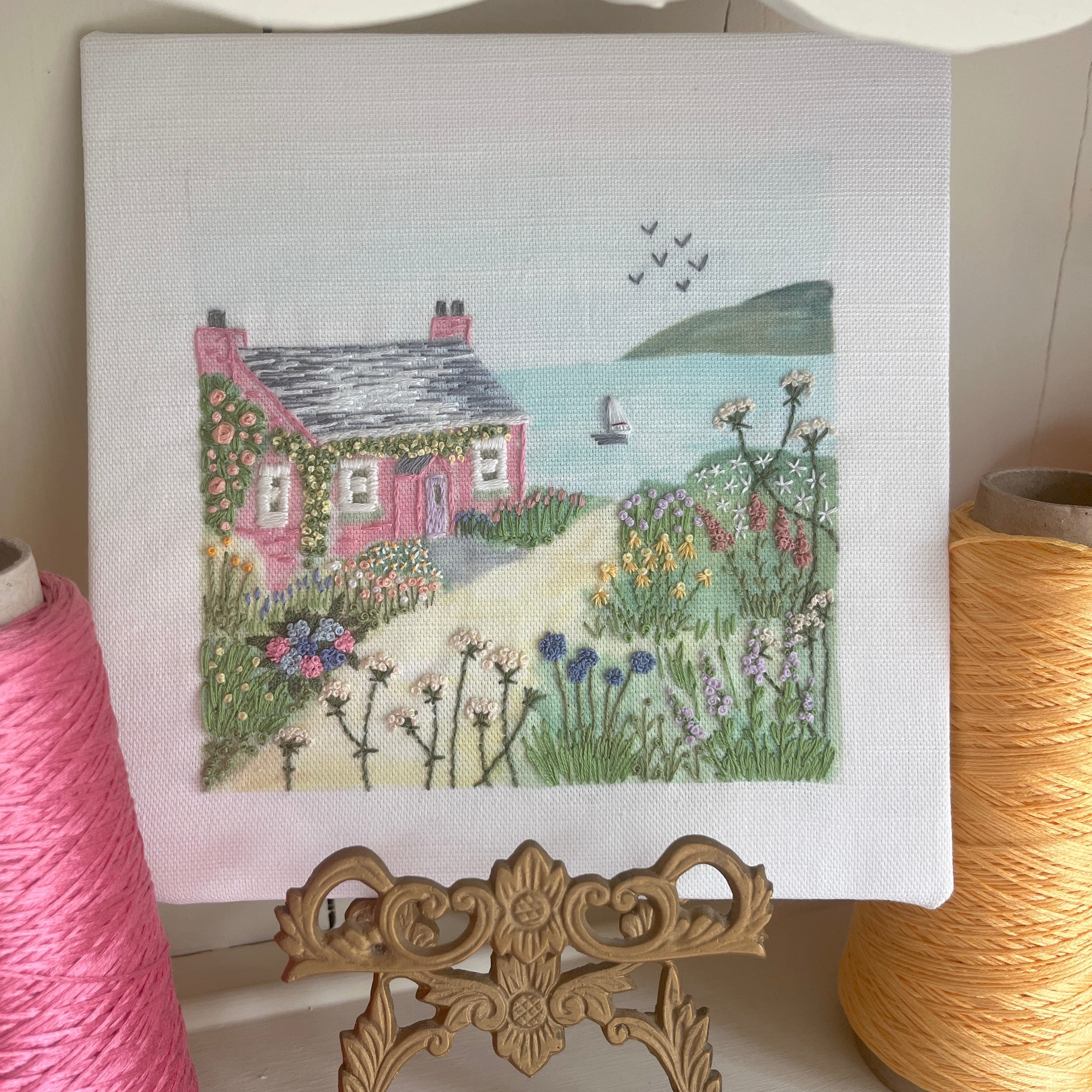 Embroidery of a pink house with a garden on a white background, surrounded by spools of thread.