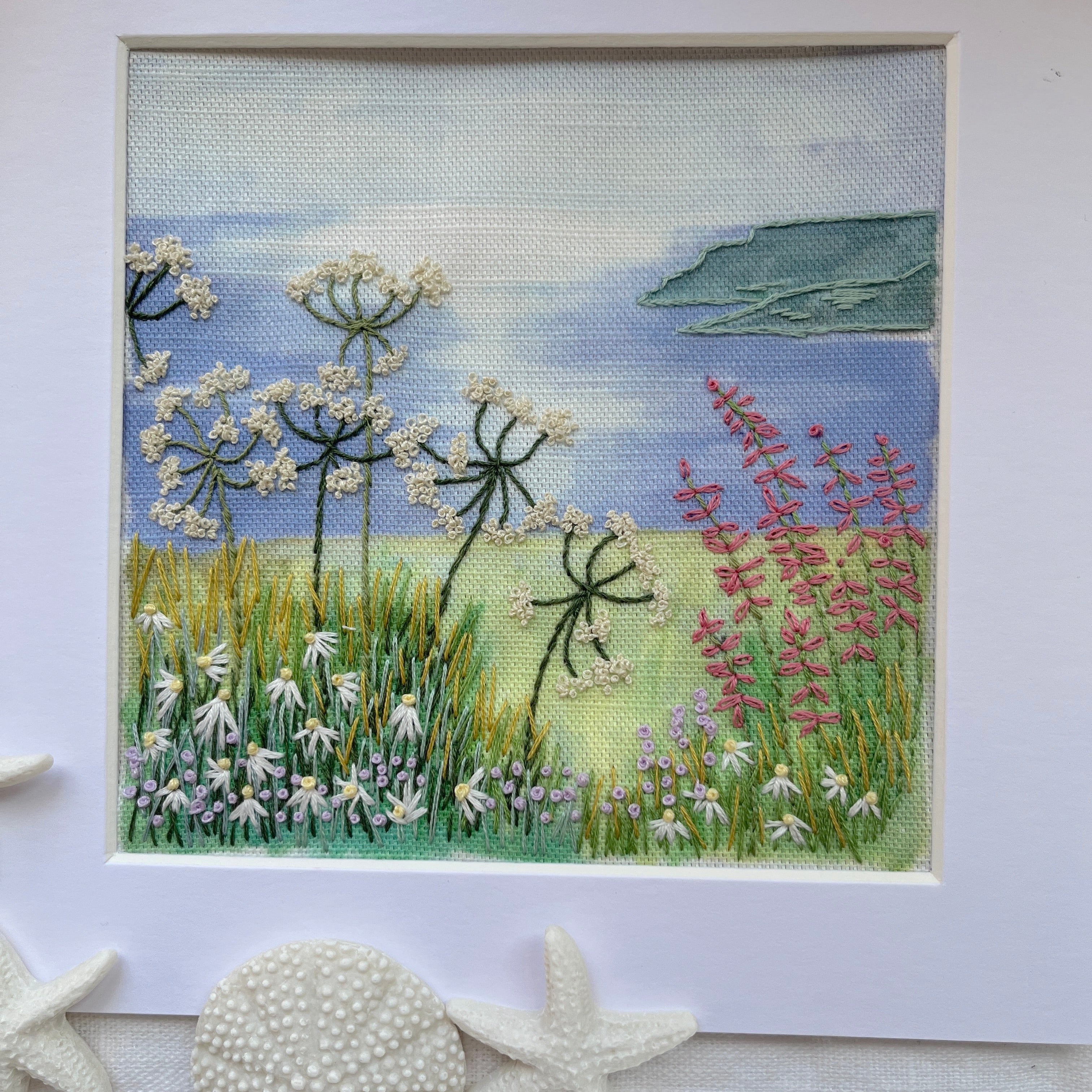 Embroidery of a field with flowers and water, framed on a white background.