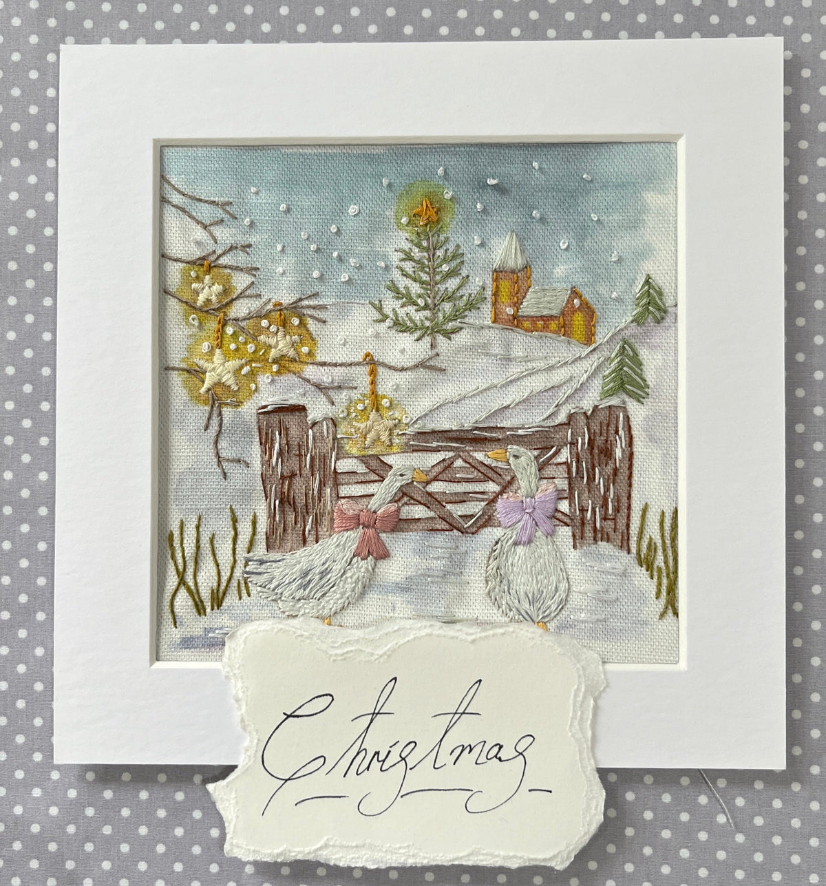 A square framed embroidered Christmas card featuring a snowy winter landscape with two white ducks, a wooden fence, trees, and a small house. A torn white paper tag at the bottom reads “Christmas” in cursive.