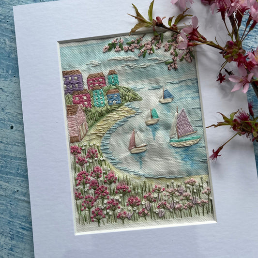 Embroidered scene of a village by a river with boats, framed on a textured surface with cherry blossoms.