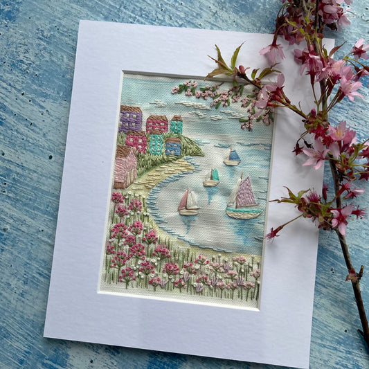 Embroidered scene of a village by a river with sailboats, framed with cherry blossoms on a textured blue surface.