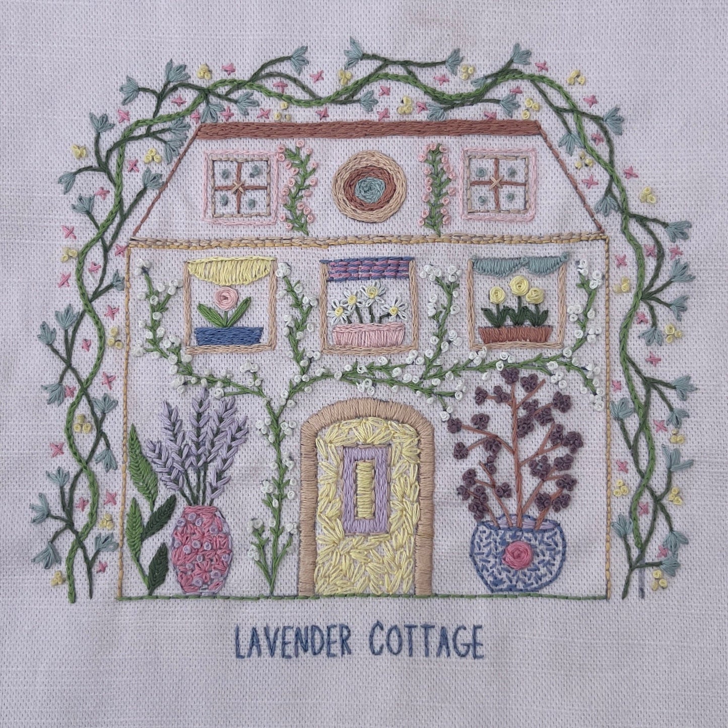 A square embroidery design on white fabric depicting a cottage labeled 'LAVENDER COTTAGE,' surrounded by green vines and flowers, with lavender and potted plants in front. The text 'LAVENDER COTTAGE' is stitched below the house.