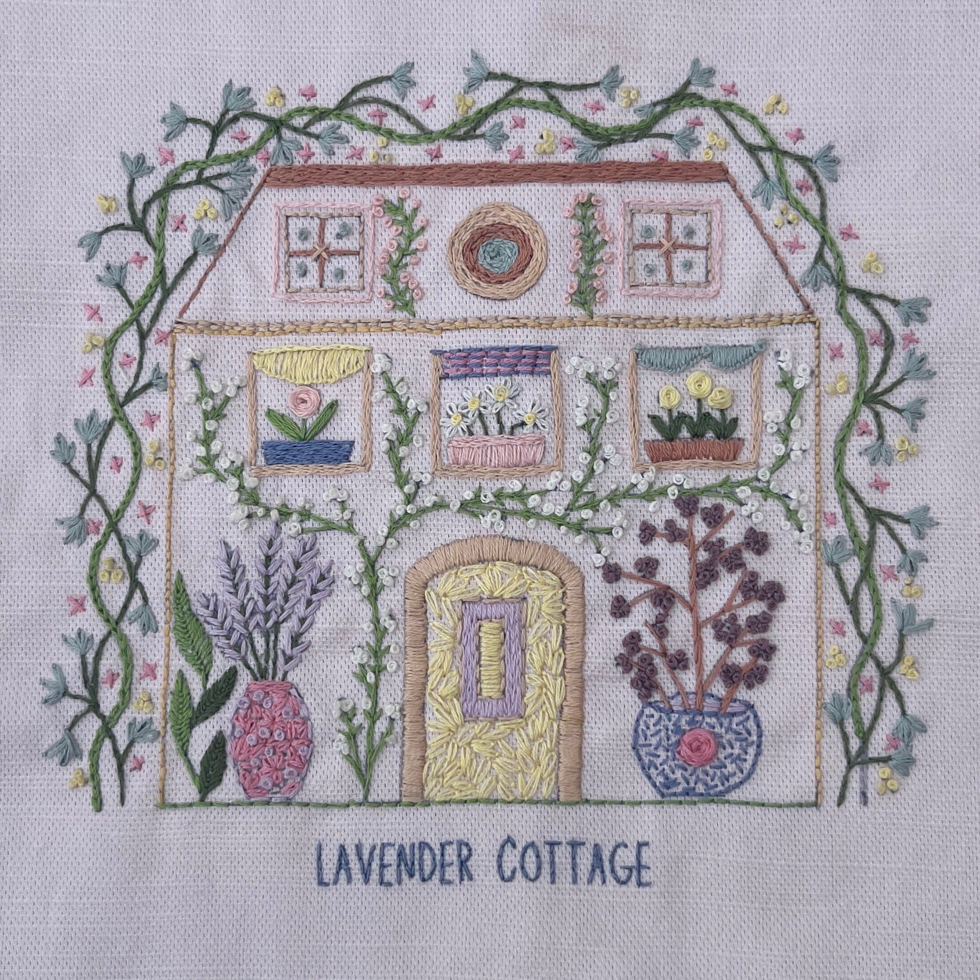 A square embroidery design on white fabric depicting a cottage labeled 'LAVENDER COTTAGE,' surrounded by green vines and flowers, with lavender and potted plants in front. The text 'LAVENDER COTTAGE' is stitched below the house.