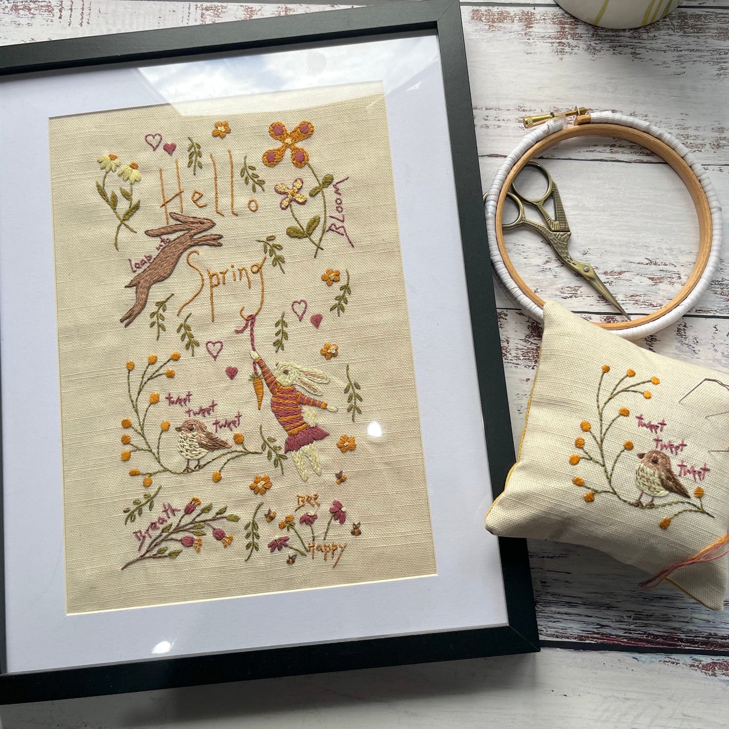 Framed embroidered artwork with 'Hello Spring' text and decorative elements on a wooden surface.