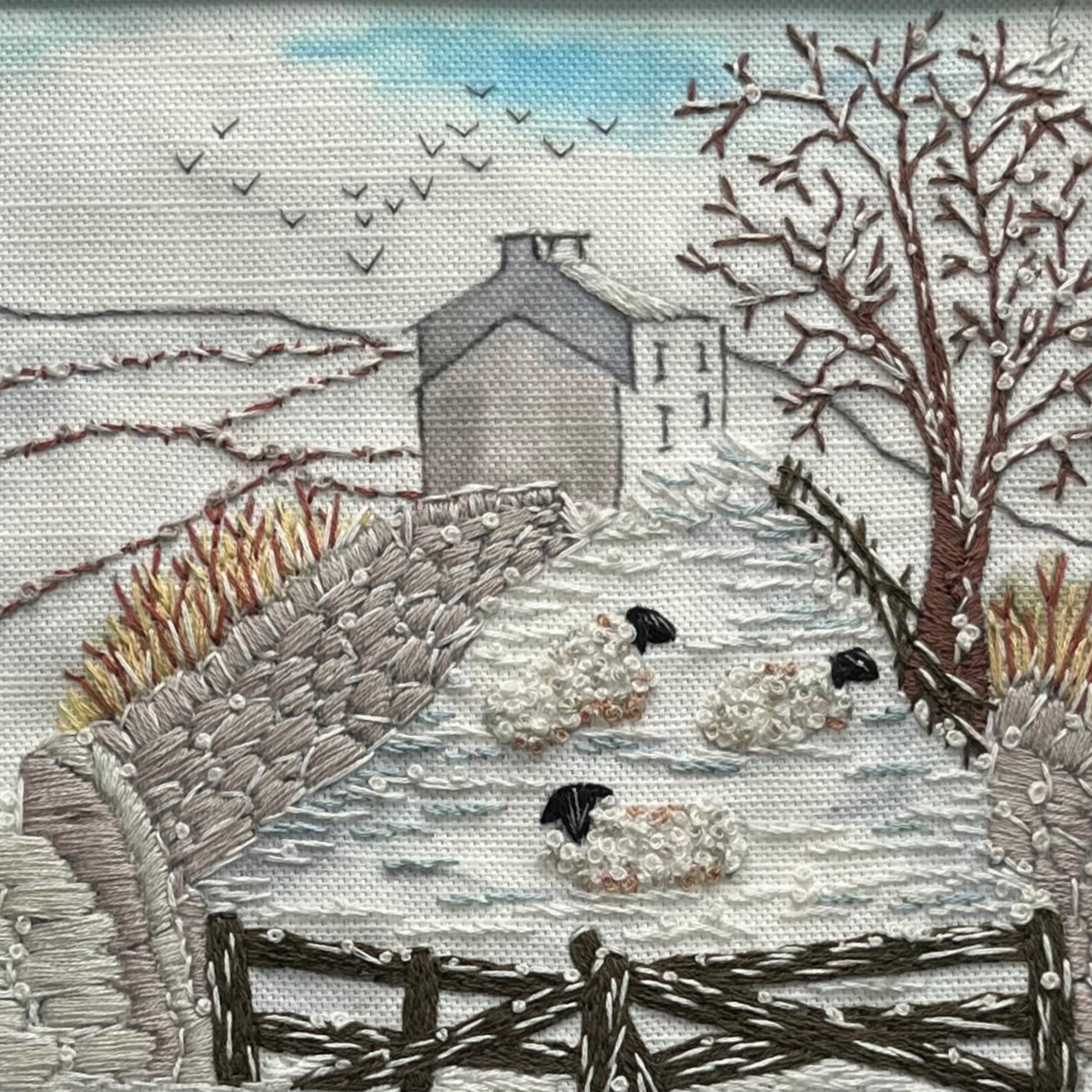 Embroidered scene of sheep in a snowy landscape with a house and trees.