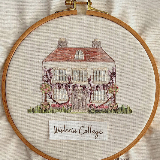 Embroidery of a house with wisteria vines in a wooden hoop on beige fabric.